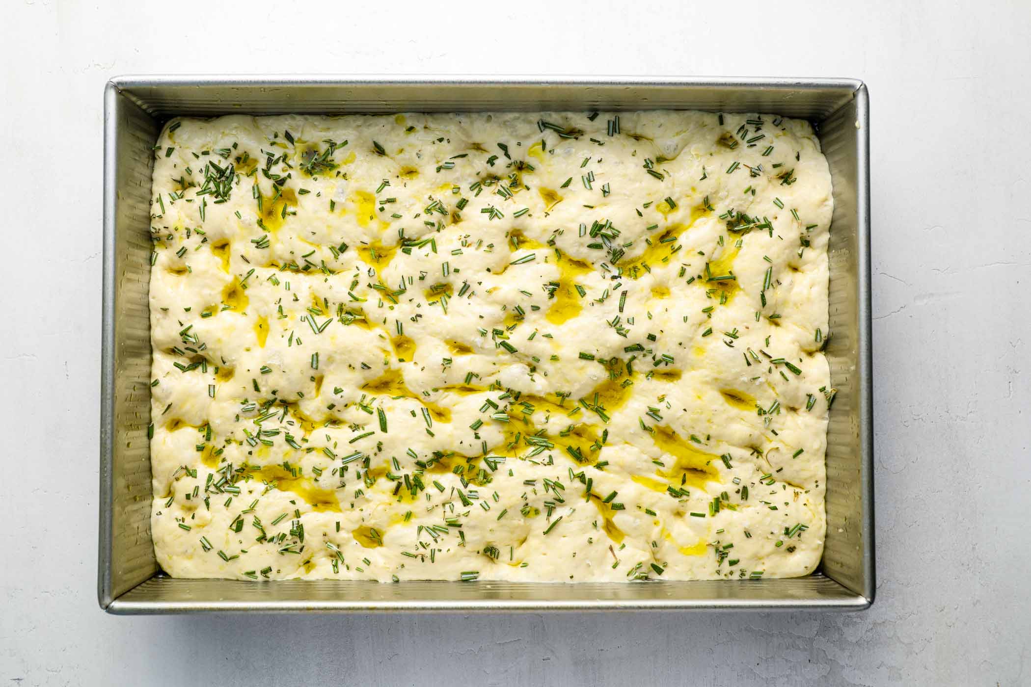 This is the only Focaccia recipe you need! It has the perfect chewy, airy texture and the best flavor. With no kneading or overnight rise required and minimal hands-on prep time, this homemade focaccia bread is incredibly fast and easy to make. We love it with rosemary and sea salt, or try one of the other flavor variations included in the recipe. Unbaked focaccia in baking pan with olive oil, rosemary and sea salt on top.